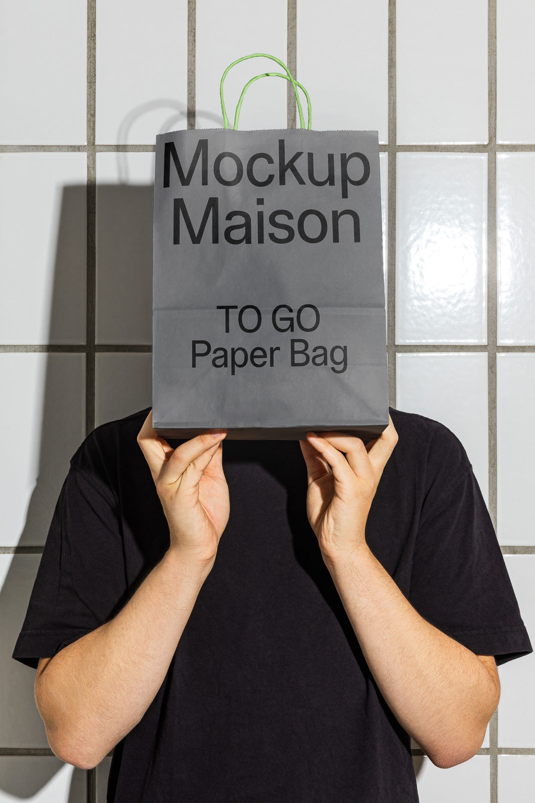 PSD mockup of a person holding a gray "Mockup Maison TO GO Paper Bag" in front of their face, with green handles, against a tiled wall backdrop.
