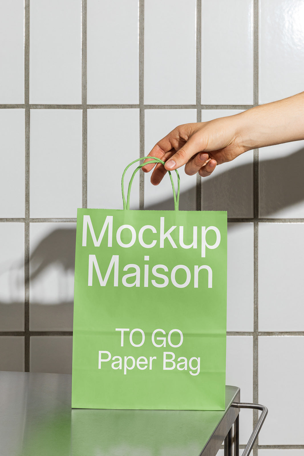 PSD mockup of a hand holding a green paper bag labeled "Mockup Maison TO GO Paper Bag," in front of a tiled wall.