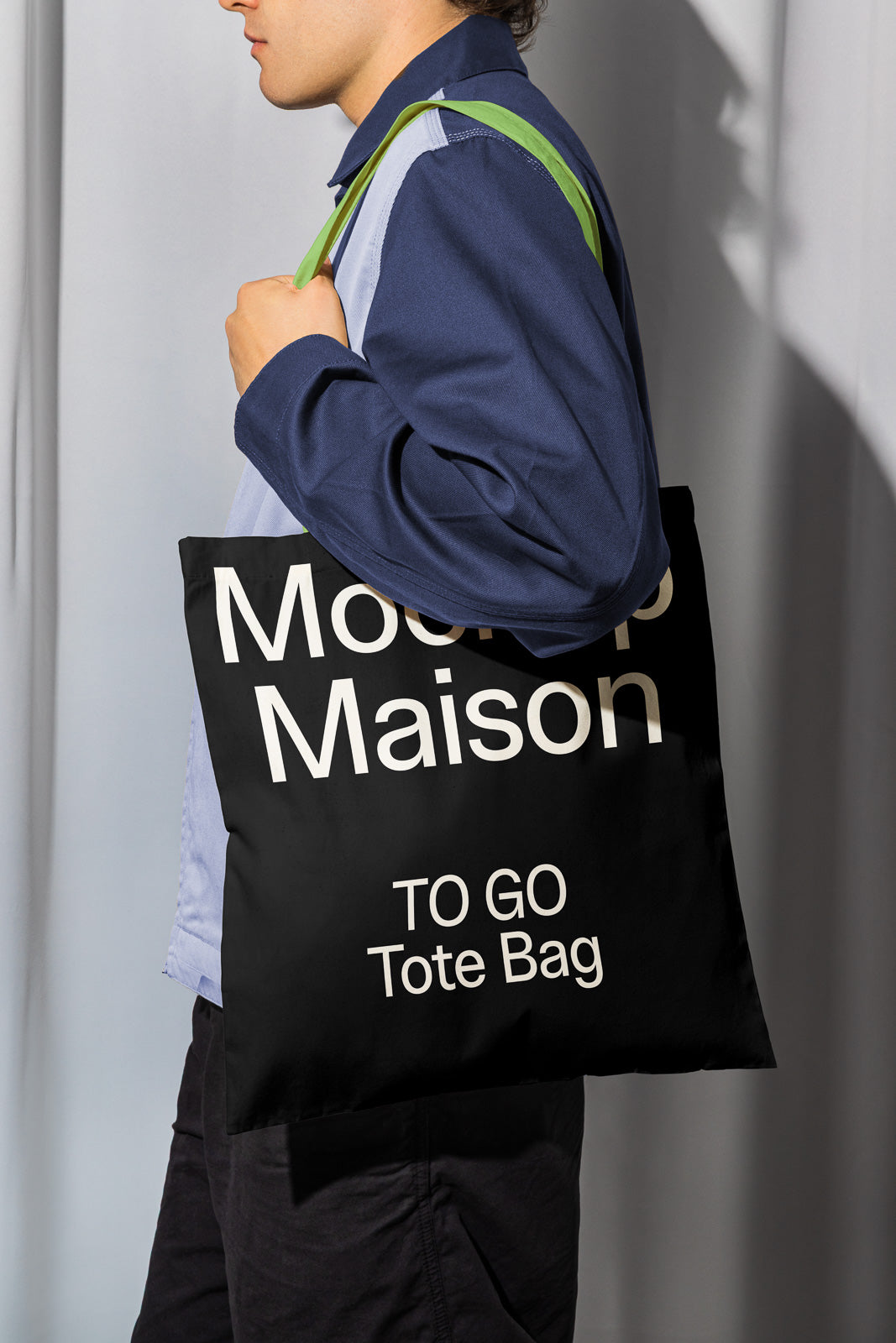 PSD mockup of a person holding a black tote bag with bold white text reading "Mockup Maison TO GO Tote Bag," draped over their shoulder.