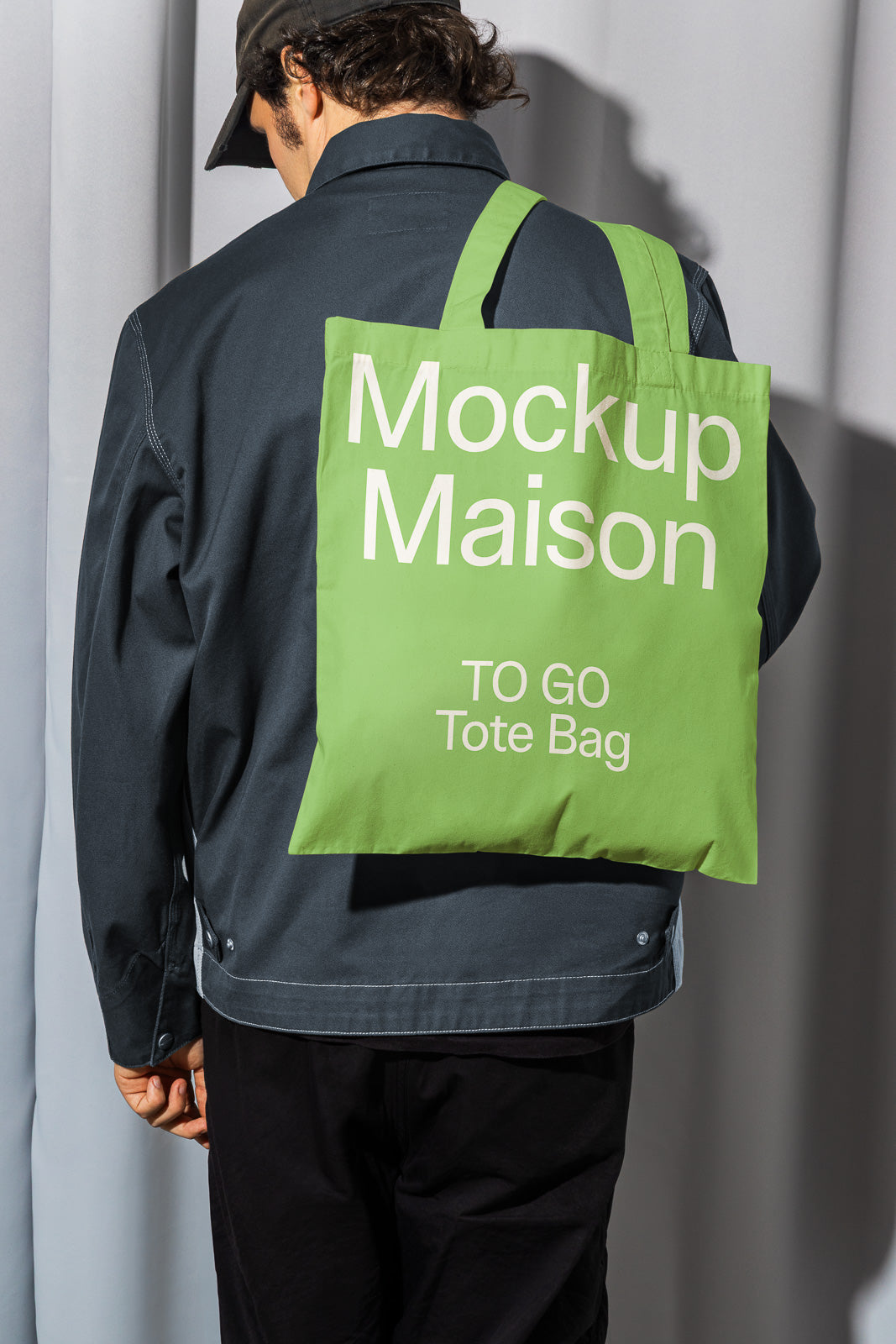 PSD mockup of a person wearing a green tote bag over their shoulder with the text "Mockup Maison TO GO Tote Bag," contrasted against a dark jacket.