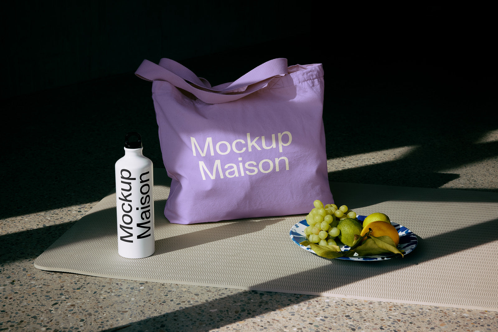 PSD mockup of a lavender tote bag labeled "Mockup Maison" near a white water bottle with the same label, and a plate of fruit.