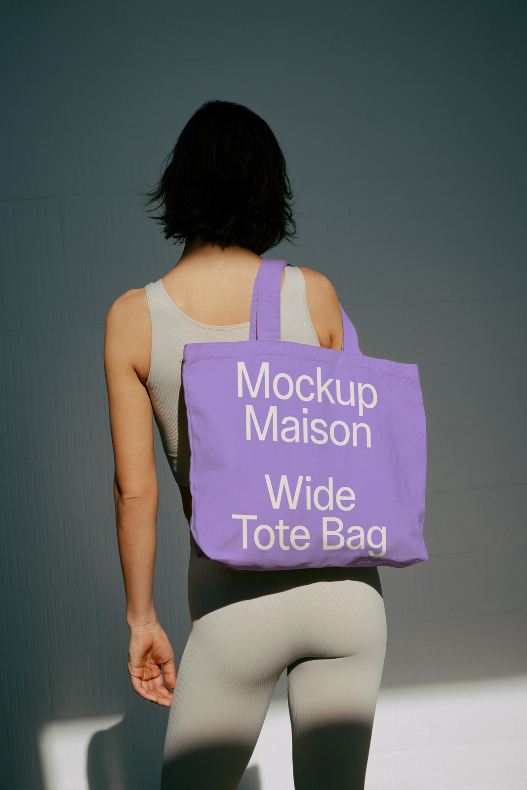PSD mockup of woman carrying a purple tote bag with "Mockup Maison Wide Tote Bag" text, wearing a light beige outfit, against a dim background.