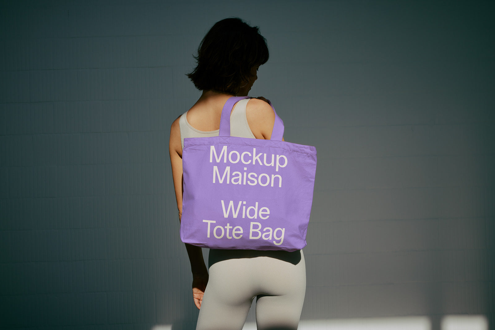 PSD mockup of a purple wide tote bag with "Mockup Maison Wide Tote Bag" printed in large white letters, carried by a person in a light attire.