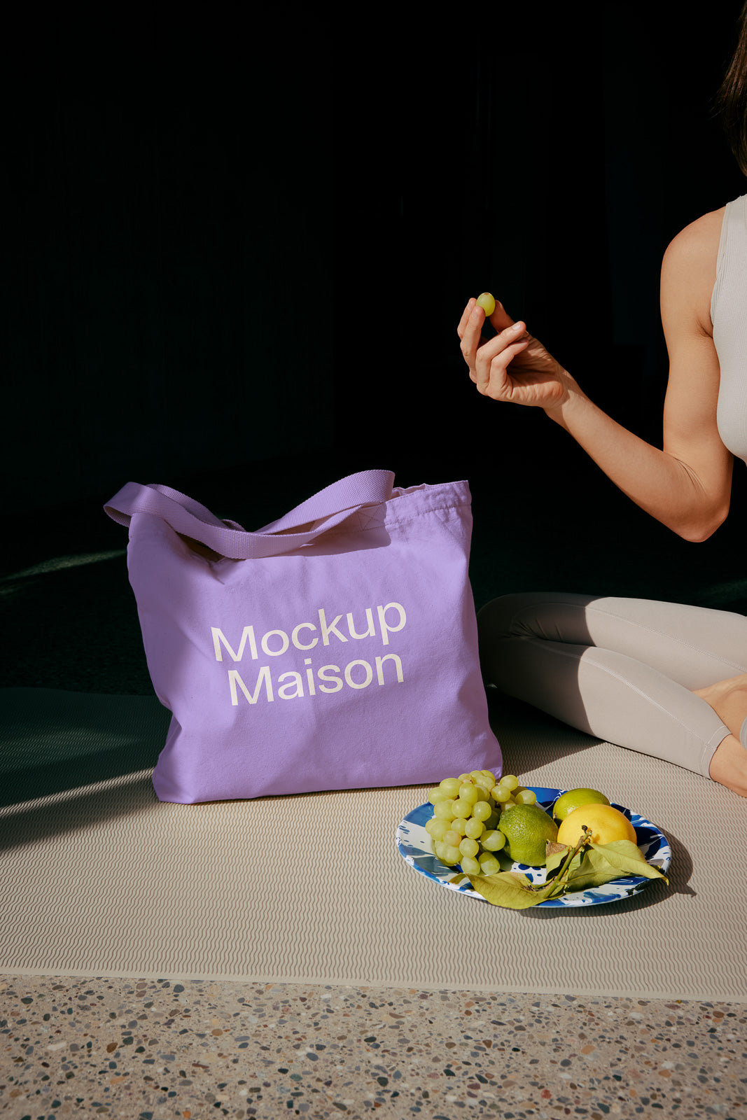 PSD mockup of a lavender tote bag with "Mockup Maison" text, placed on a mat beside a plate of green grapes and citrus fruits with a person holding a grape.