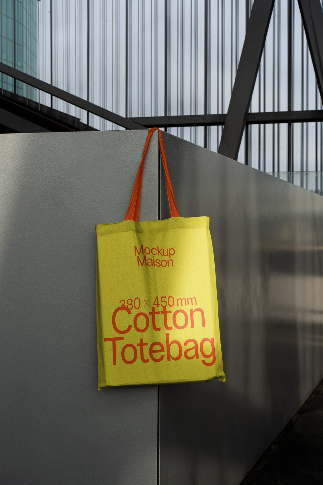 PSD mockup of a yellow cotton tote bag with orange handles hanging on a gray metal surface, displaying text: "Mockup Maison, 380 x 450 mm".