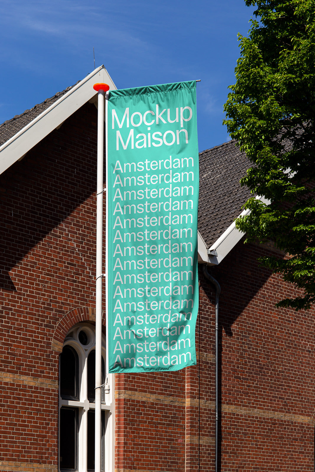 PSD mockup of vertical flag mounted on pole, featuring repeated text "Amsterdam" under a clear blue sky against a brick building backdrop.