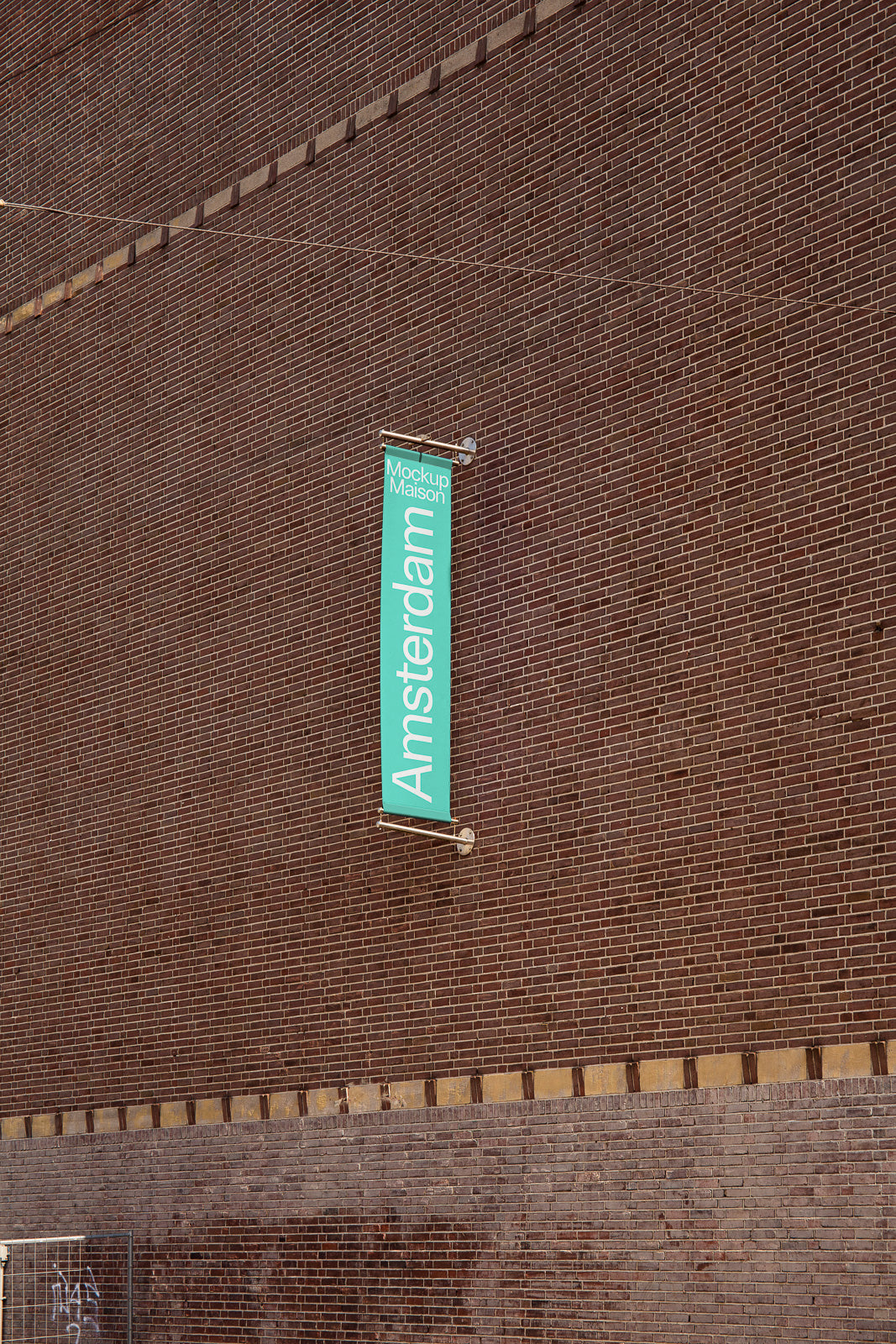 PSD mockup of a green vertical banner on a brown brick wall with the text “Amsterdam.” The banner is attached to metal rods and casts a shadow.