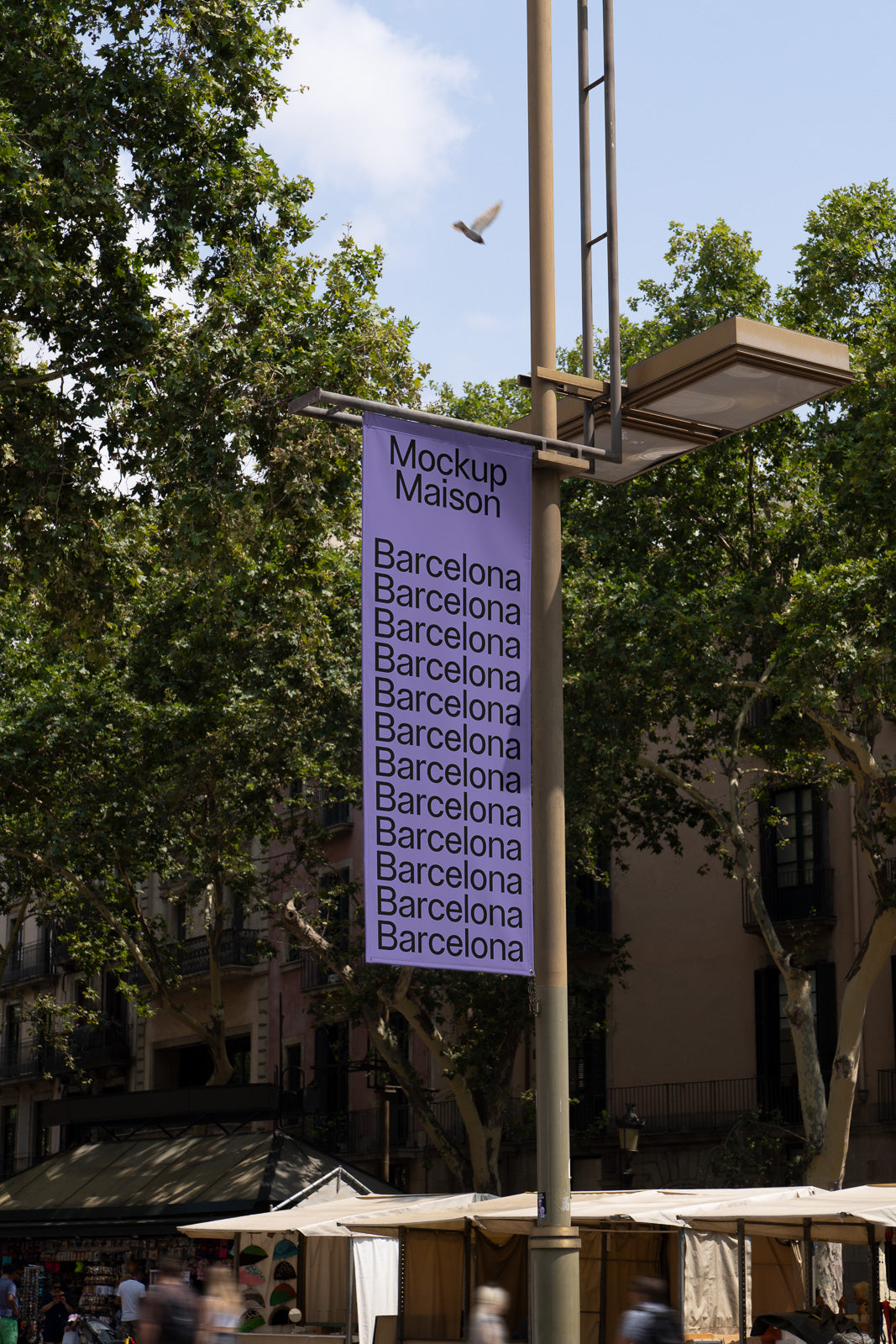 PSD mockup of a street banner hanging from a pole, featuring the words "Mockup Maison Barcelona" repeated. Trees and buildings visible in background.