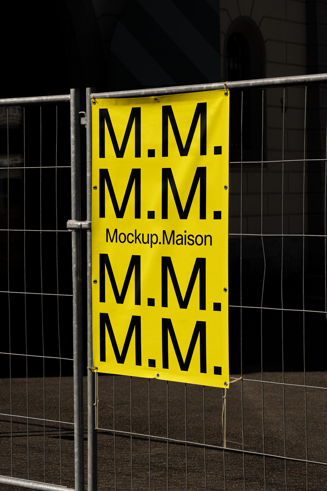 PSD mockup of a yellow banner with "M.M." repeated and "Mockup.Maison" printed, attached to a metal fence against a dark background.