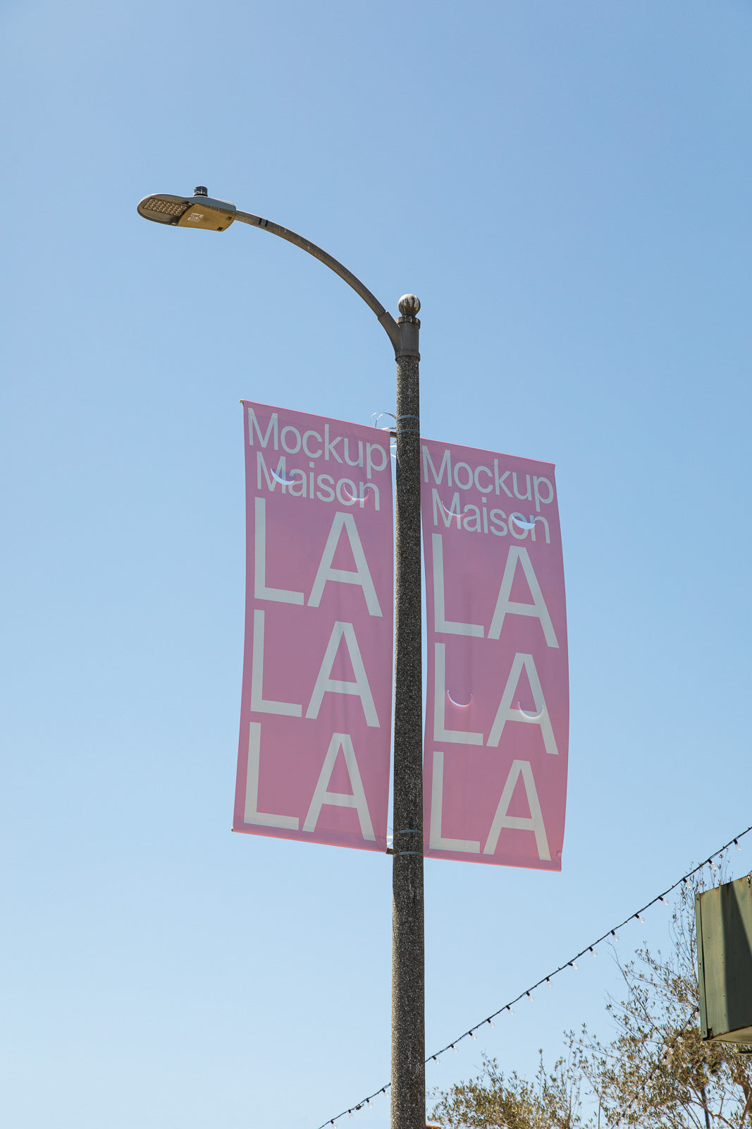 PSD mockup of a streetlight displaying pink banners with the text "Mockup Maison LA LA LA" against a clear blue sky.