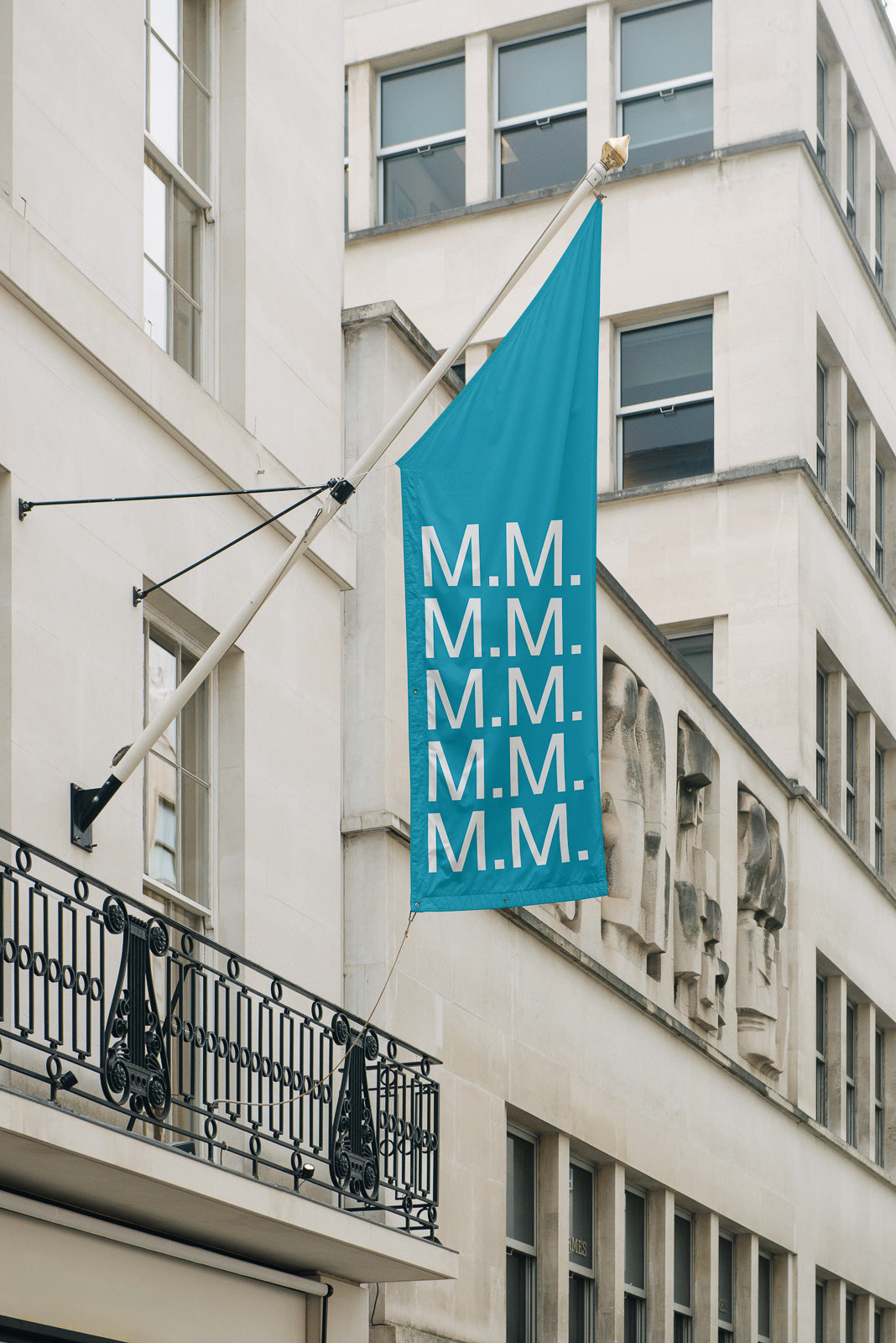 PSD mockup of a vertical teal flag with repeated "M.M." text, hanging from a building facade with ornate iron railing and stone architecture.