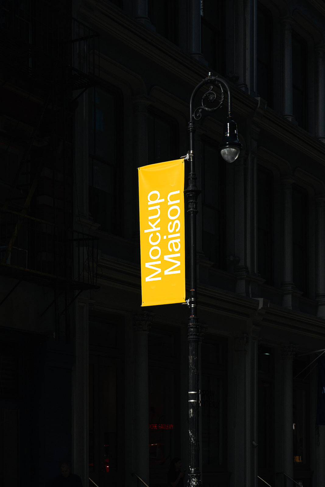 PSD mockup of a vertical yellow street banner hanging on a decorative metal pole, with the words "Mockup Maison" against a dark building background.