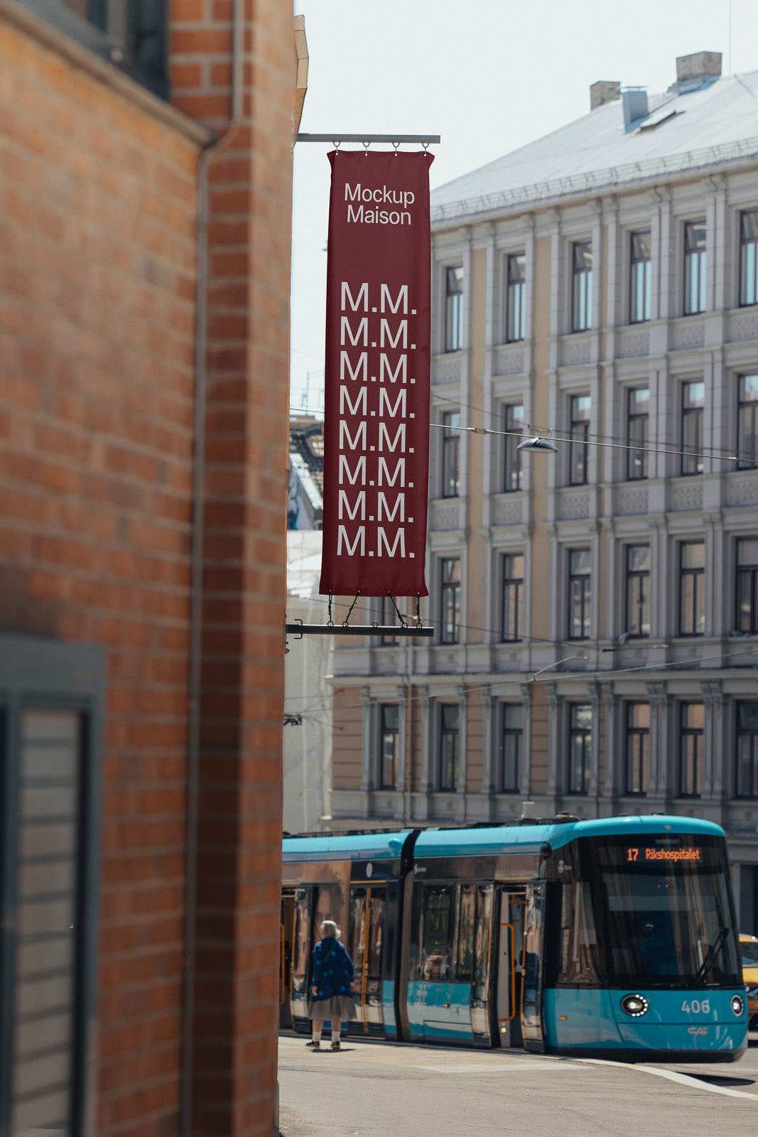 PSD mockup of a vertical street banner with repeating initials "M.M." displayed on a red background, positioned above a city street with a tram passing by.
