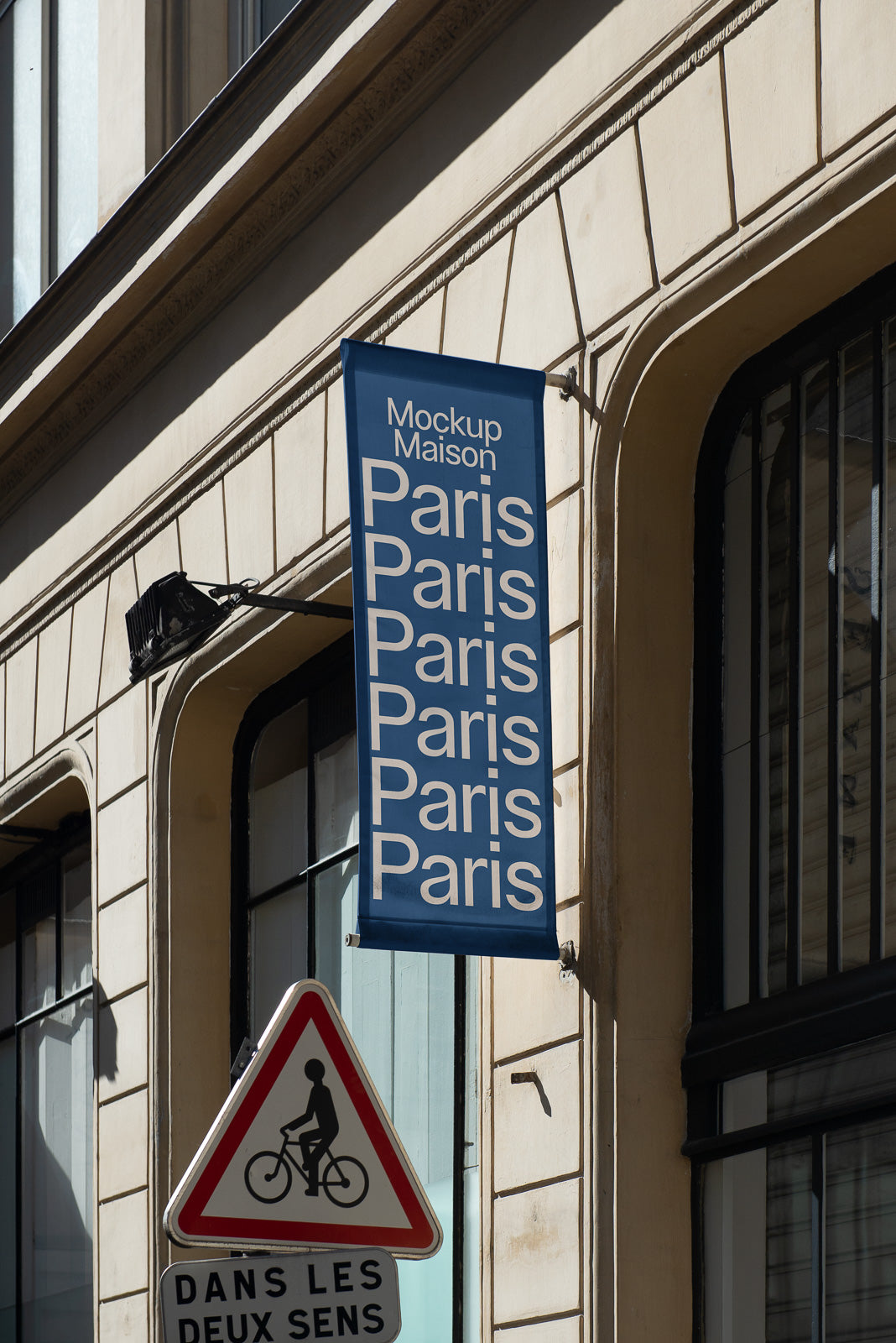 PSD mockup of a vertical blue banner on a building facade with repeating text "Paris" and "Mockup Maison" at the top, next to a bicycle traffic sign.