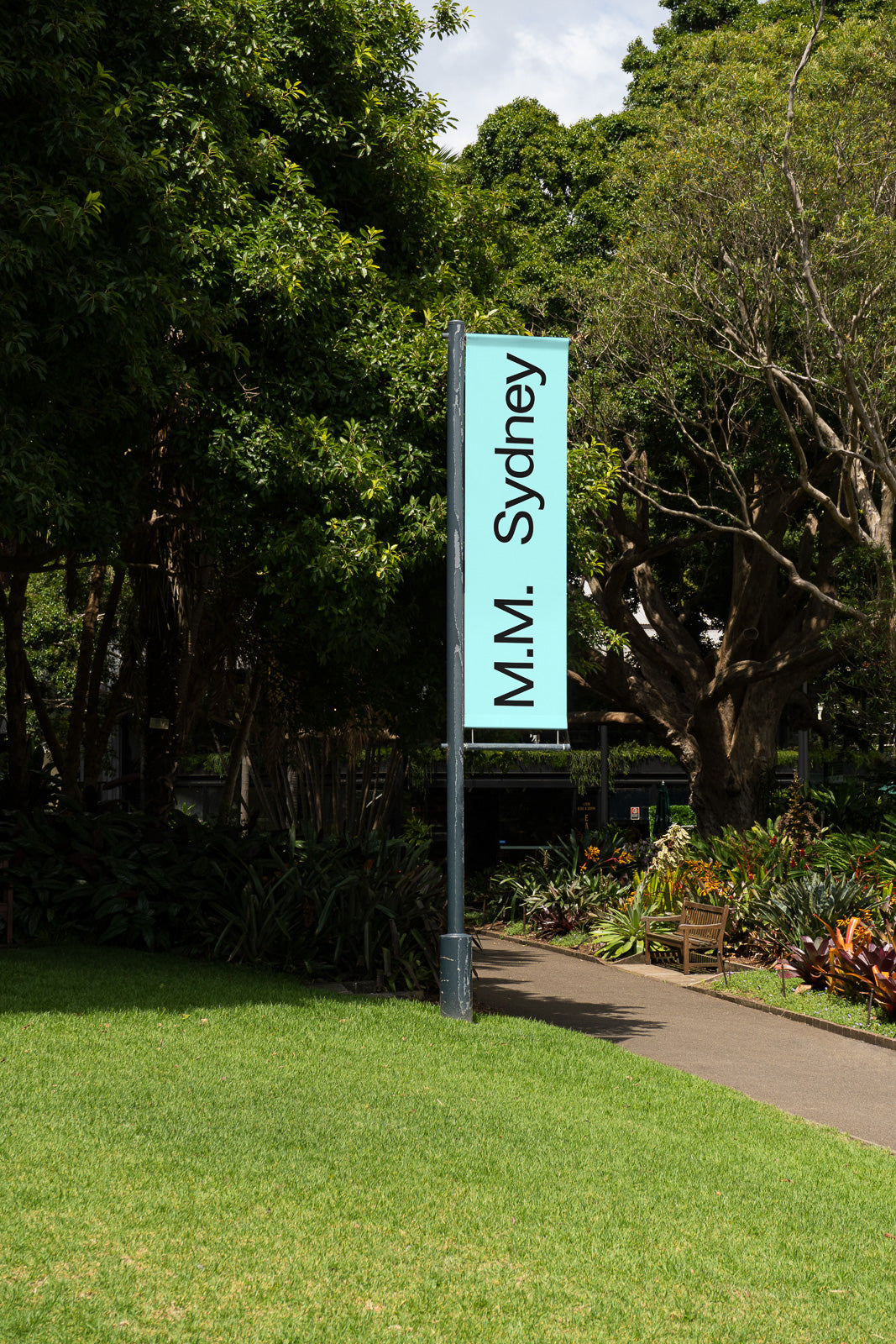 PSD mockup of a vertical banner on a pole in a park setting, with lush greenery and a walking path, featuring the text "M.M. Sydney" on a turquoise background.