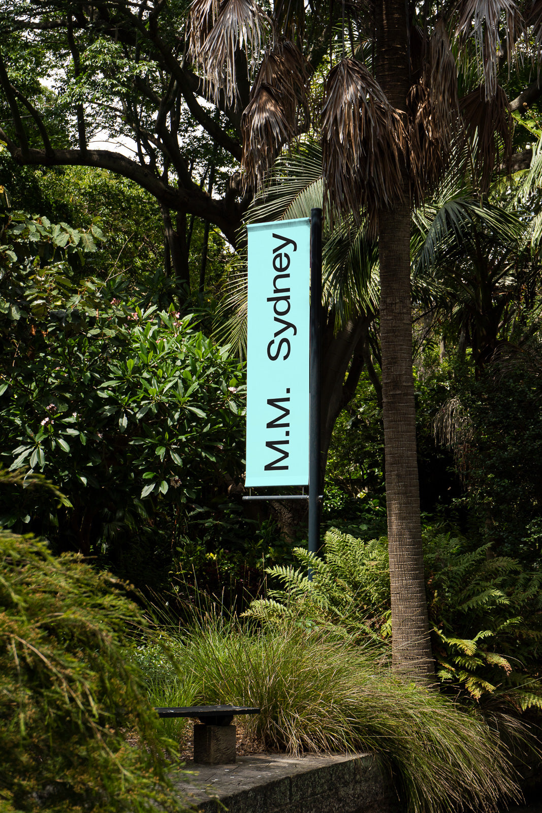 PSD mockup of a vertical banner displaying "M.M. Sydney" mounted on a pole amidst lush greenery and trees, with a stone bench nearby.
