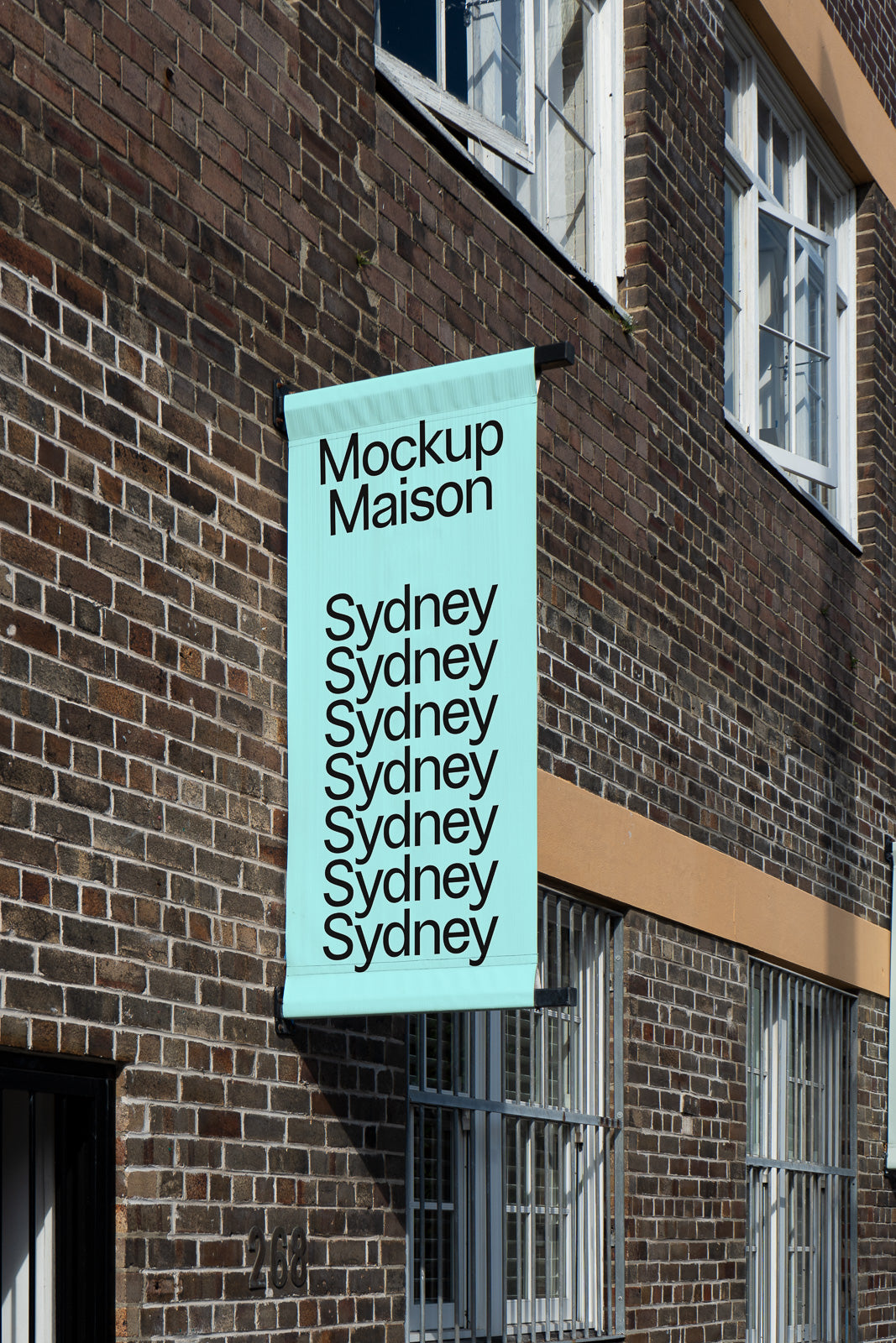 PSD mockup of a vertical banner hanging on a brick building facade, displaying "Mockup Maison" and "Sydney" text repeated.