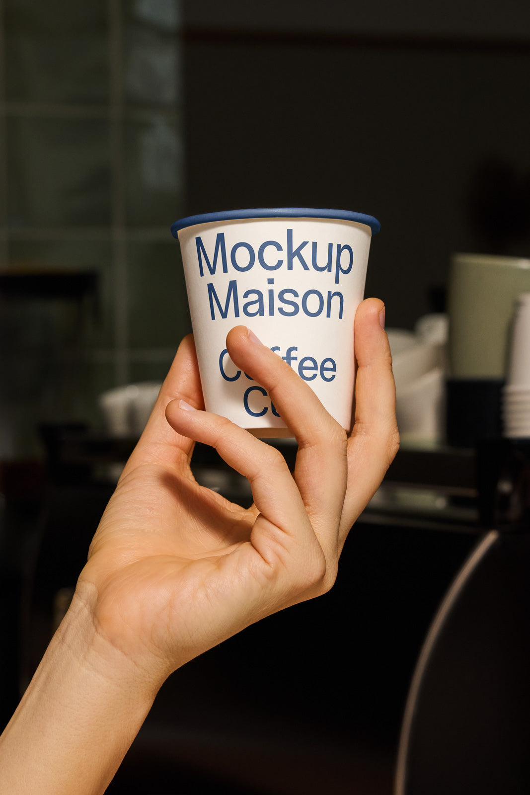 PSD mockup of a hand holding a paper coffee cup labeled "Mockup Maison Coffee" with a blurred background in a cafe setting.