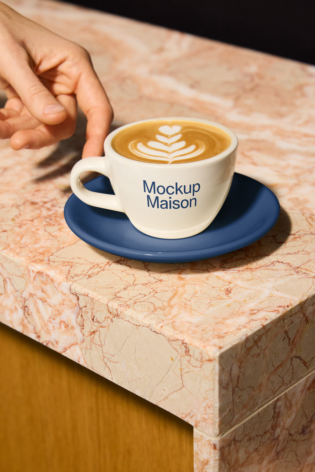 PSD mockup of a white coffee cup with latte art on a blue saucer, placed on a marble surface as a hand reaches for it.