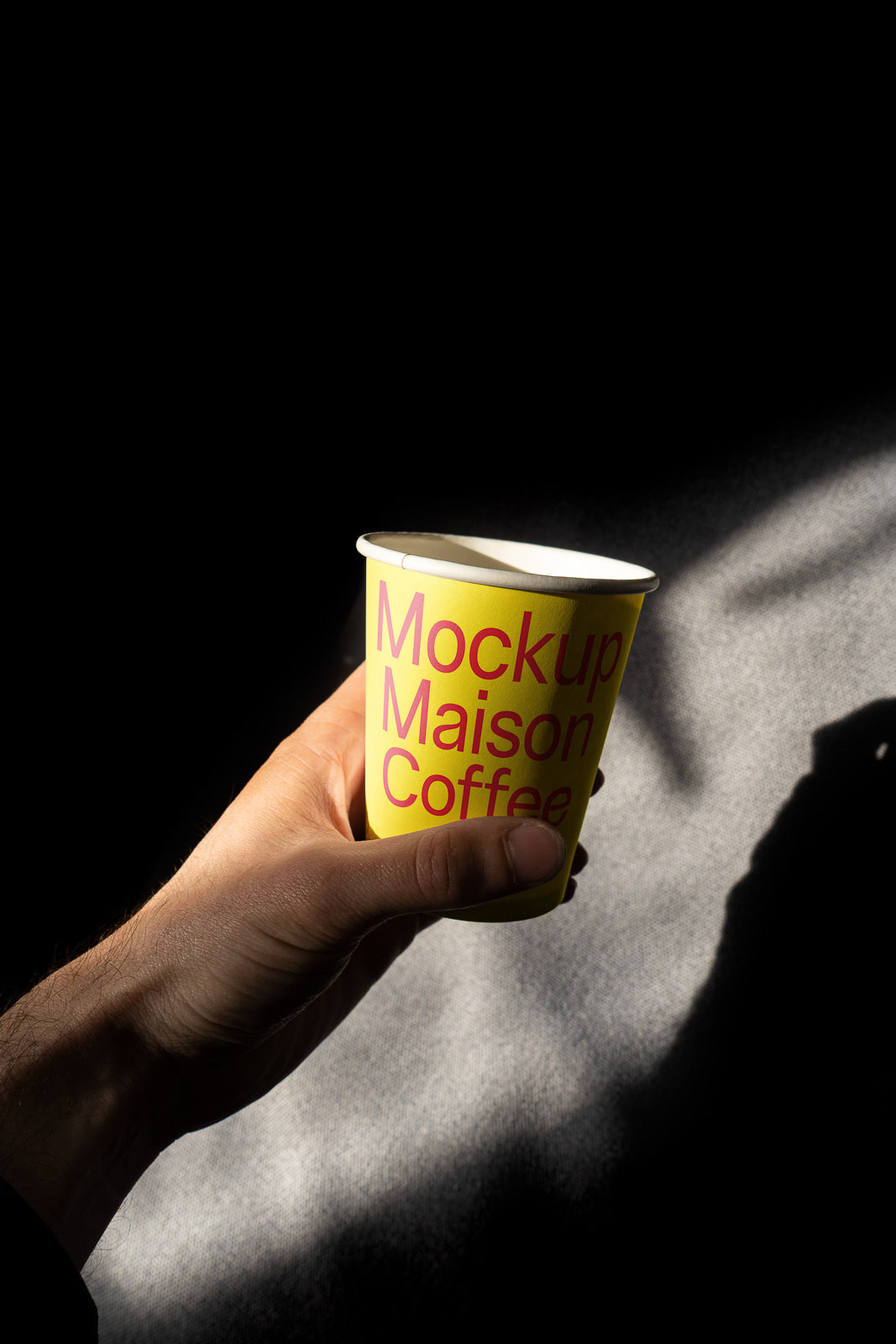 PSD mockup of a hand holding a yellow coffee cup labeled "Mockup Maison Coffee" against a dark background, with light casting shadows.