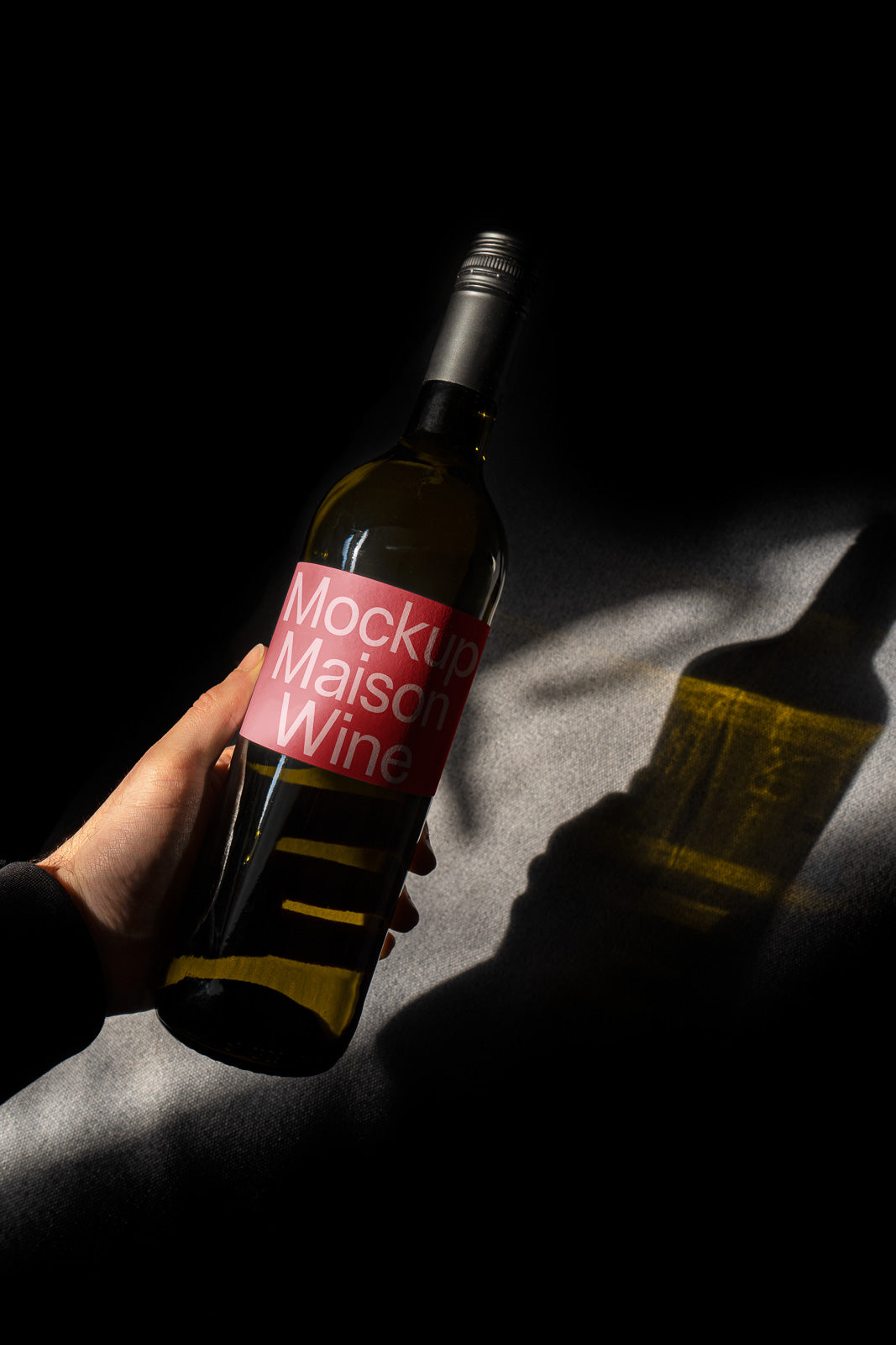 PSD mockup of a wine bottle with a pink label held in a hand, casting a shadow on a textured surface under dramatic lighting.