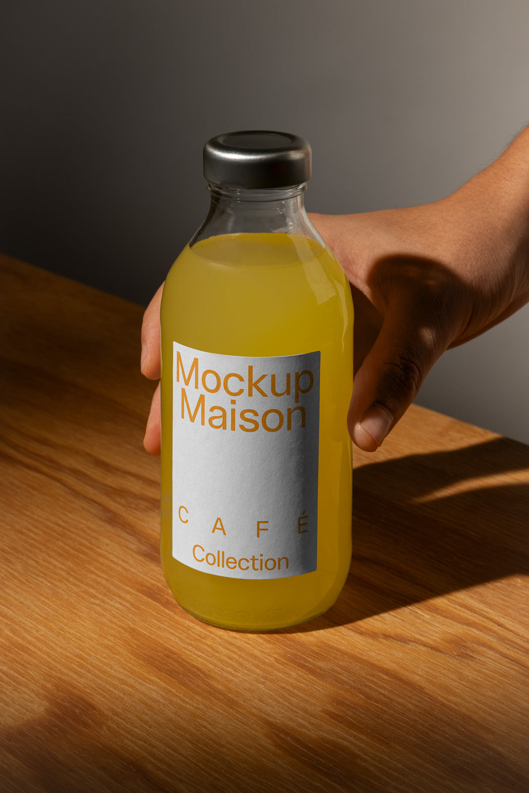 PSD mockup of a hand holding a bottle with a sleek metal cap, featuring a label with text "Mockup Maison" and "Café Collection" on a wooden table.