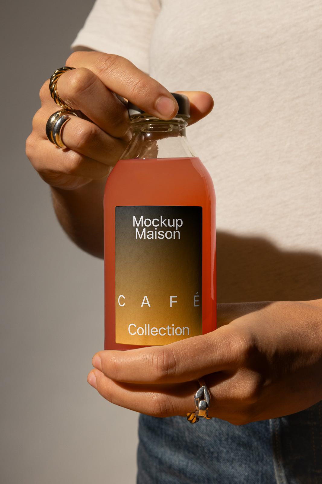 PSD mockup of a person holding a clear glass bottle filled with orange liquid, labeled "Mockup Maison Café Collection," wearing rings on their fingers.