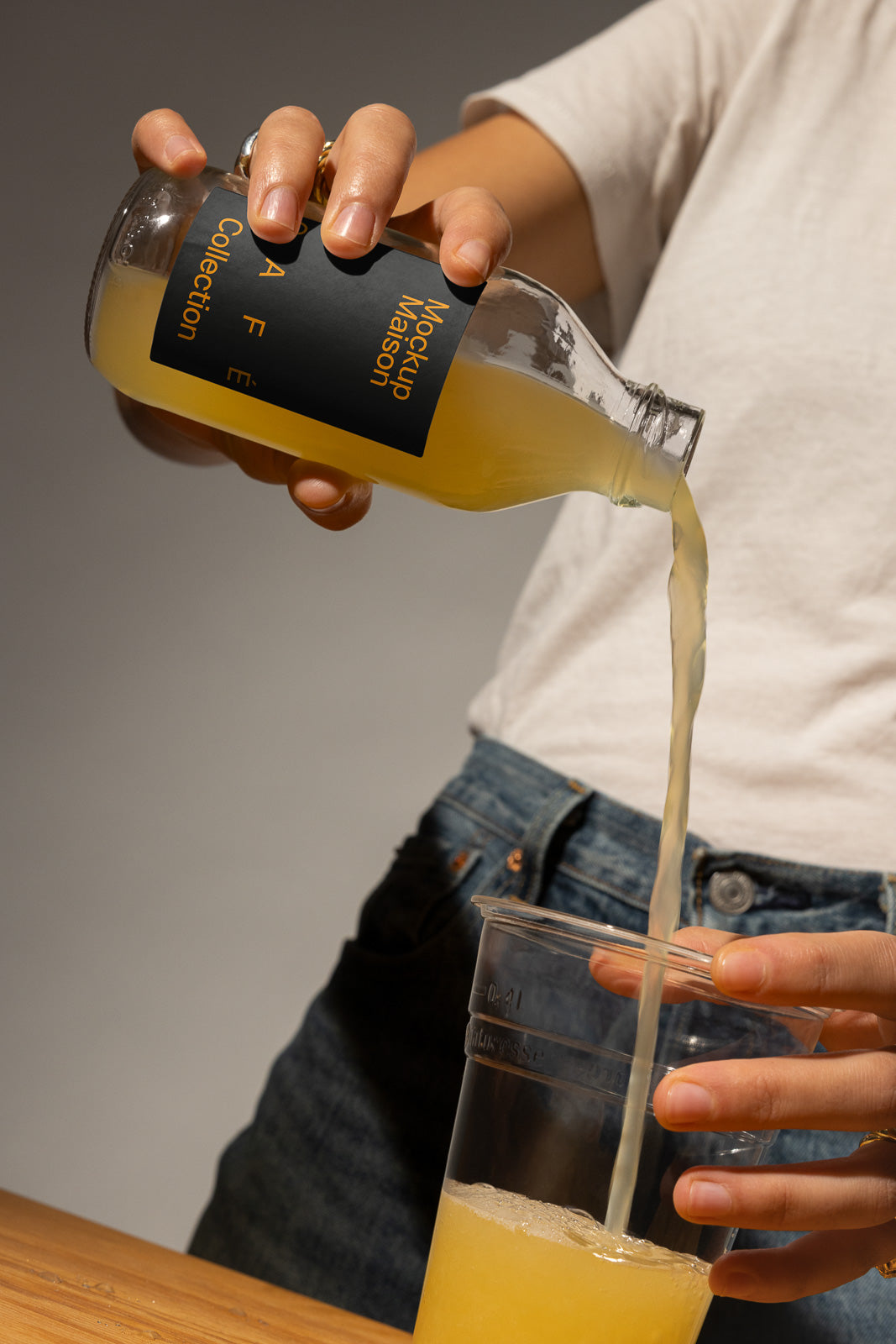 PSD mockup of a hand pouring yellow liquid from a bottle into a clear plastic cup placed on a wooden table.