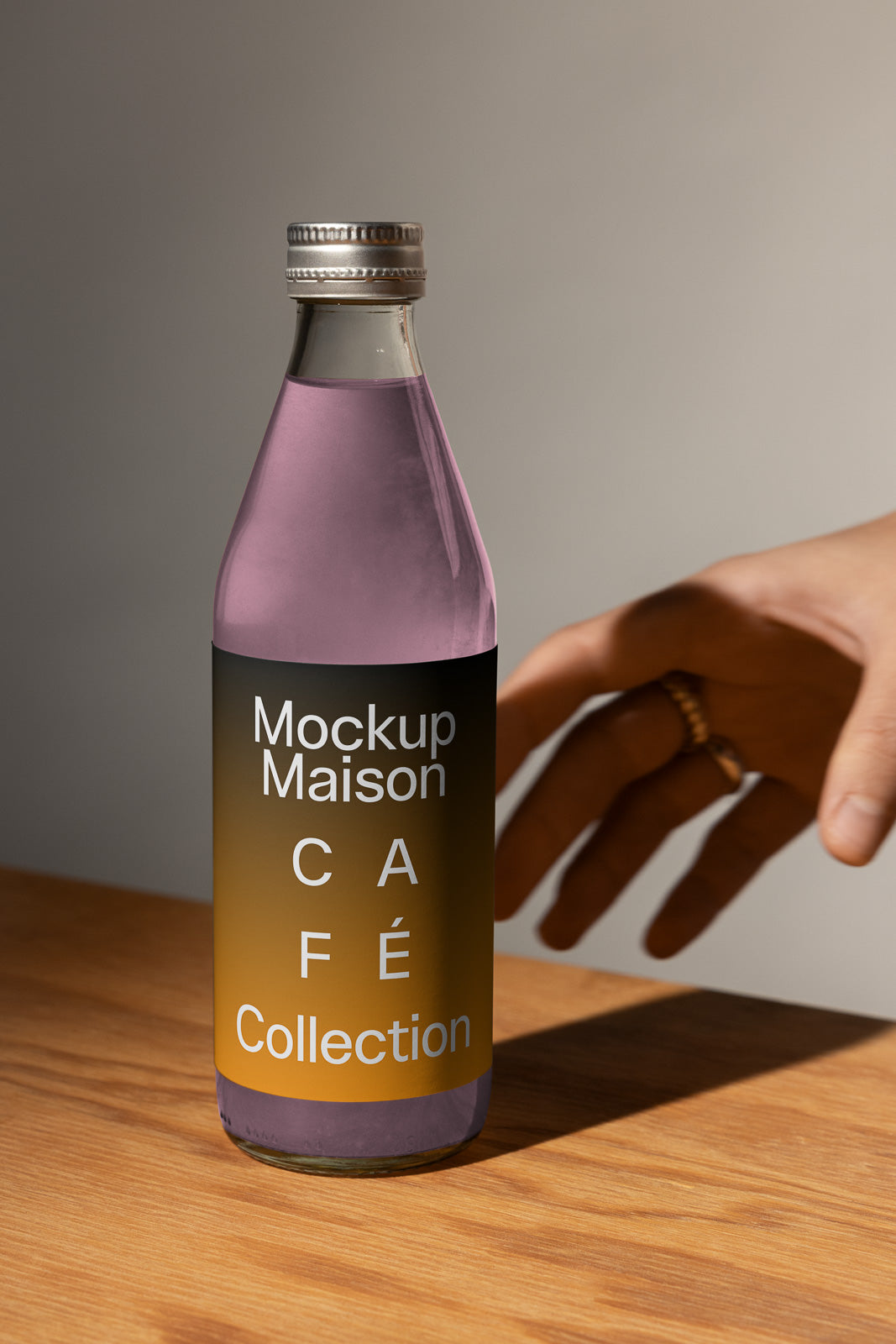 PSD mockup of a glass bottle with purple liquid on a wooden surface, featuring a label design titled "Mockup Maison Café Collection" beside a hand reaching towards it.