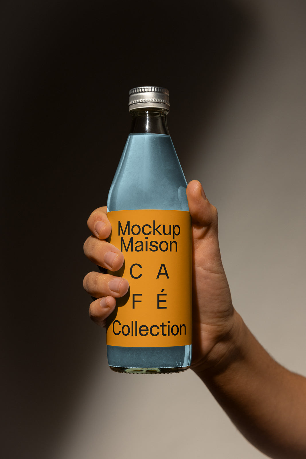 PSD mockup of a hand holding a glass bottle labeled "Mockup Maison Café Collection" with an orange label and metallic cap.