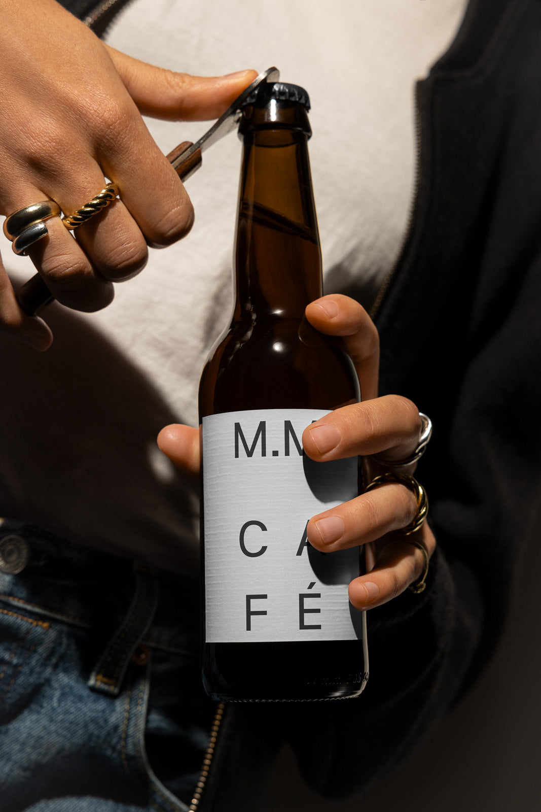 PSD mockup of a person opening a brown glass bottle with a minimalist white label that reads "M.N. CAFE," highlighting hands adorned with rings.
