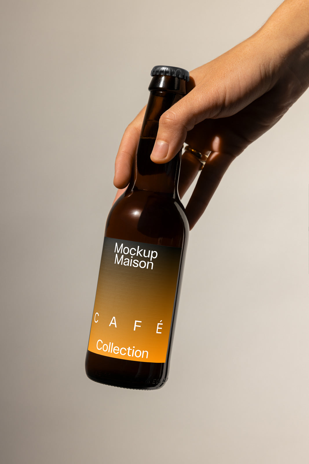 PSD mockup of a hand holding a brown bottle labeled "Mockup Maison Café Collection" with a gradient orange and brown design.
