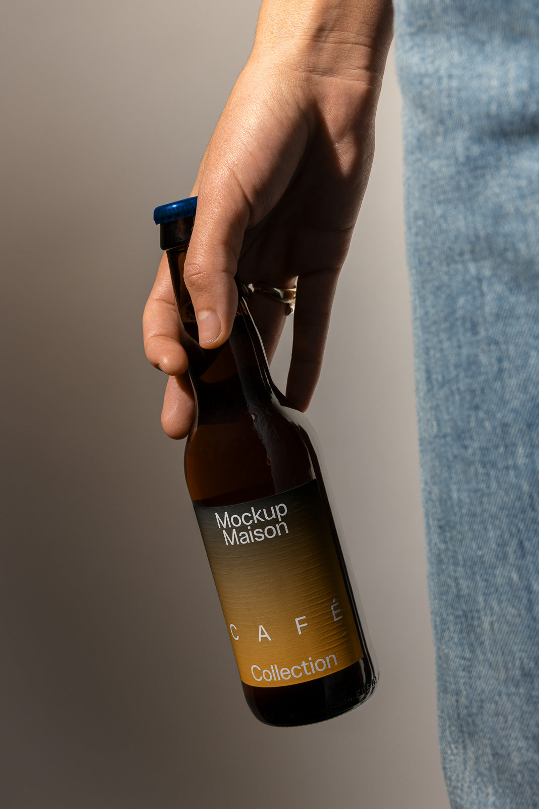 PSD mockup of a hand holding a brown bottle labeled "Mockup Maison Café Collection" with a blue cap against a neutral background.