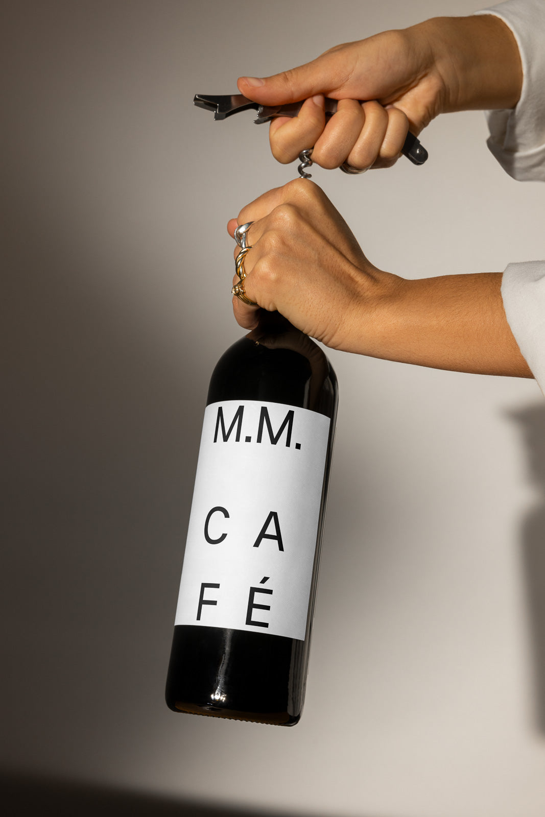 PSD mockup of hands opening a wine bottle with a corkscrew. The label on the bottle reads "M.M. CAFÉ" in bold letters, set against a neutral background.
