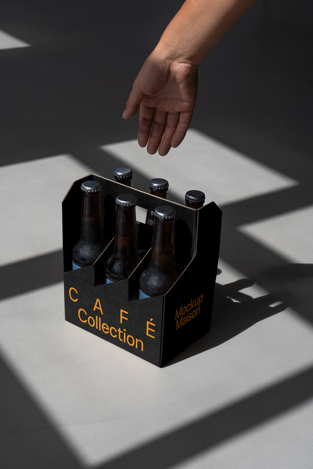 PSD mockup of a hand reaching for a six-pack of brown bottles labeled "Café Collection" in a dark carrier on a sunlit gray surface.