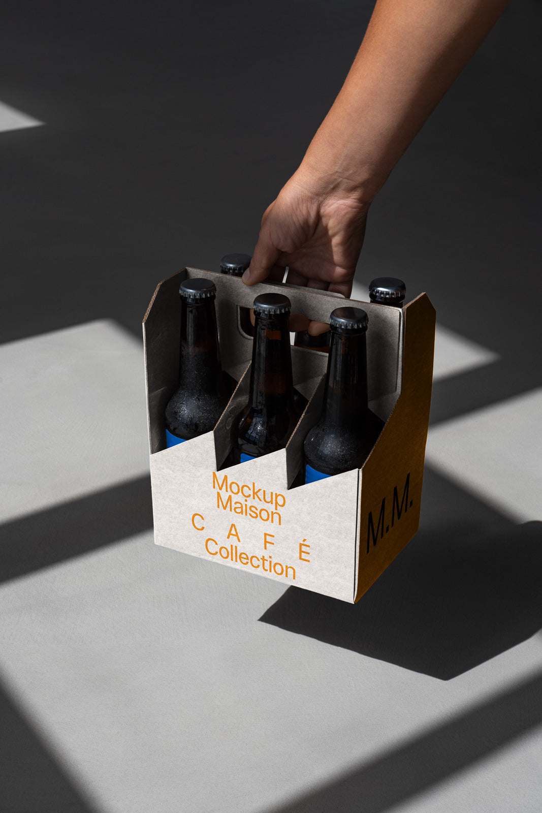 PSD mockup of a hand holding a six-pack of bottled beverages in a cardboard carrier labeled "Mockup Maison Café Collection" on a gray background with shadows.