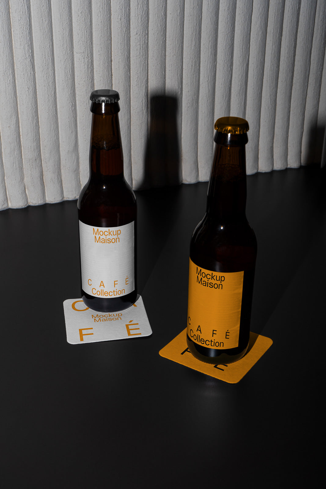 PSD mockup of two glass beer bottles with "Mockup Maison Café Collection" labels, placed on matching coasters against a white ribbed backdrop.