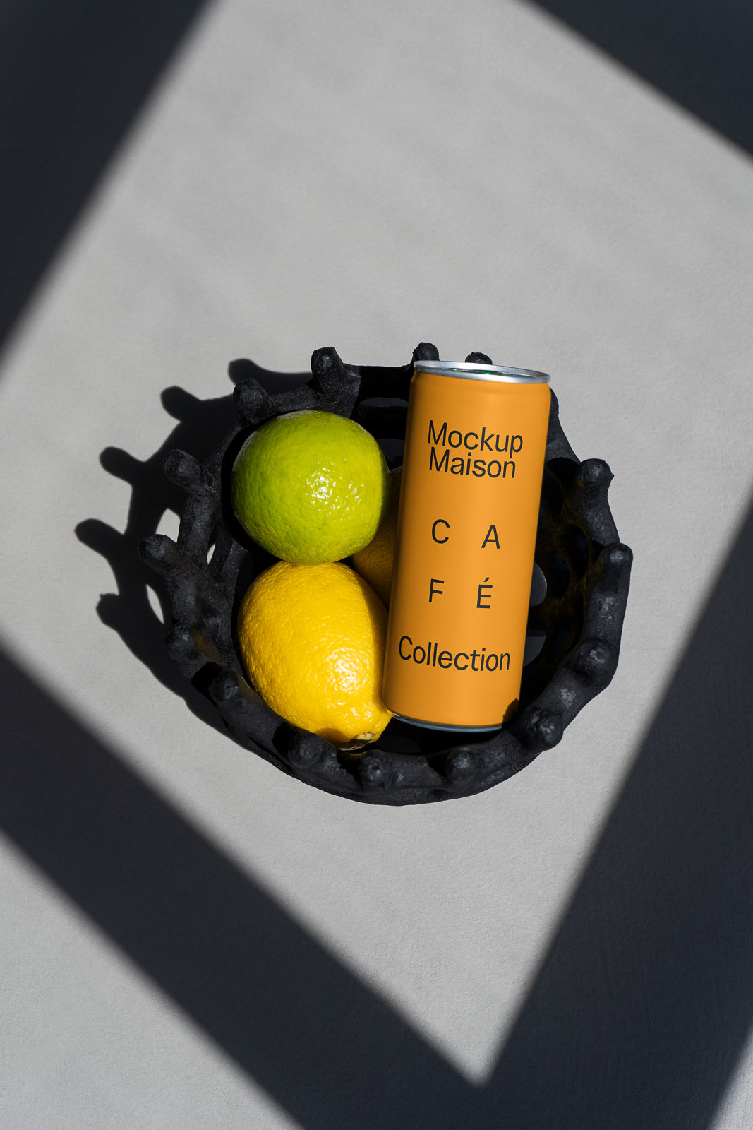 PSD mockup of a yellow can labeled "Mockup Maison Café Collection" placed in a black basket with a lime and a lemon, casting shadows on a gray surface.