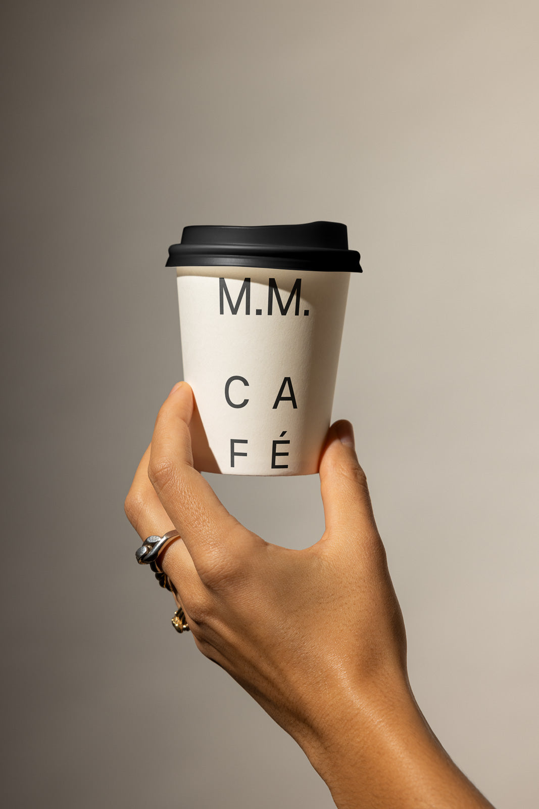 PSD mockup of a hand holding a white coffee cup with black lid, labeled "M.M. CAFÉ." The design features minimalist lettering against a neutral background.