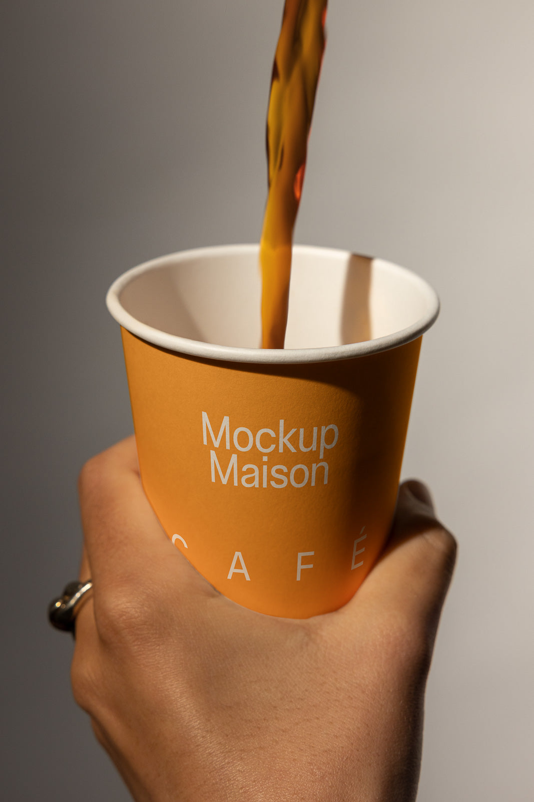PSD mockup of a yellow paper cup labeled "Mockup Maison Café" held in hand, with coffee being poured in, capturing a realistic presentation of branding.