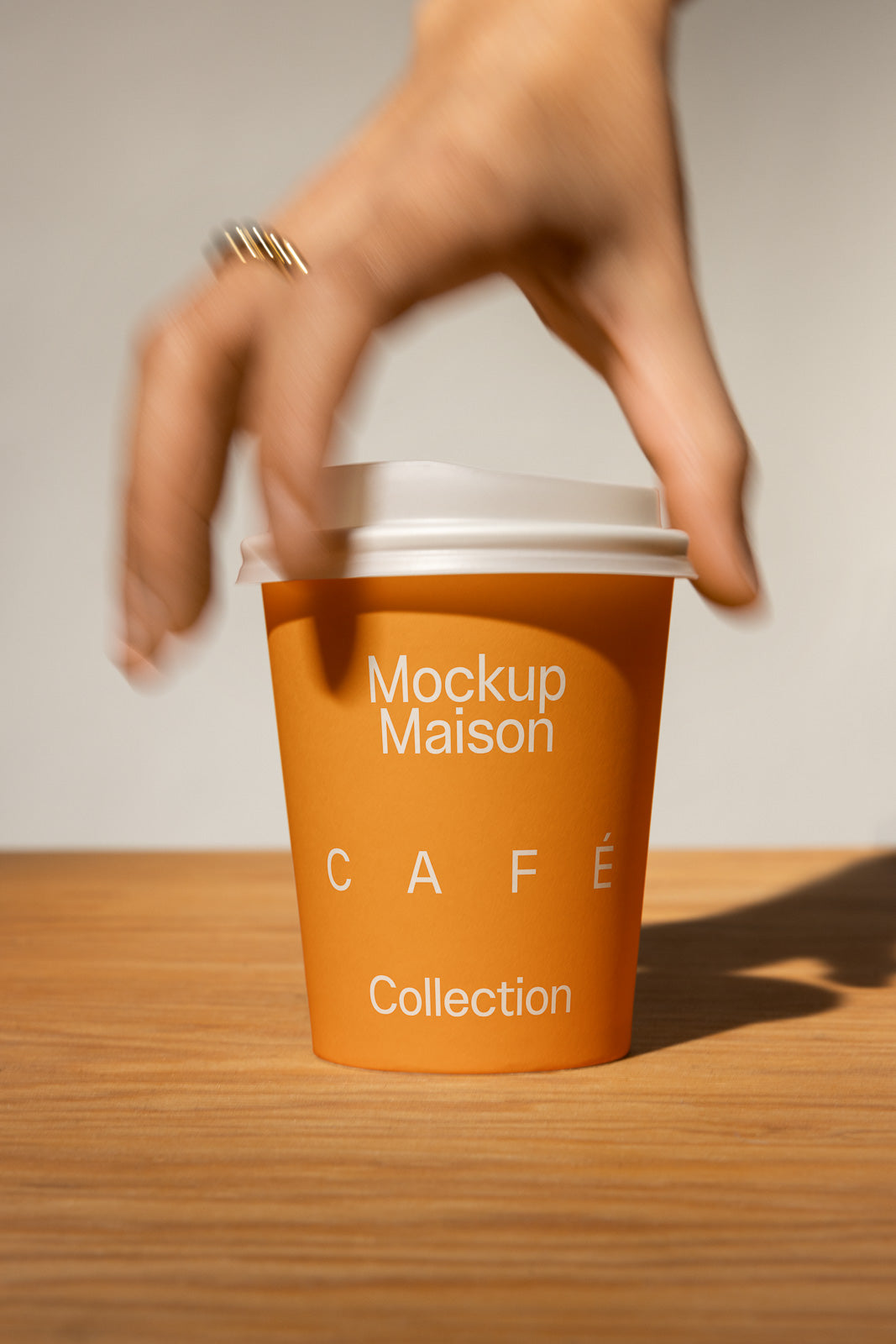 PSD mockup of an orange coffee cup labeled "Mockup Maison Café Collection" on a wooden surface with a hand reaching to pick it up.