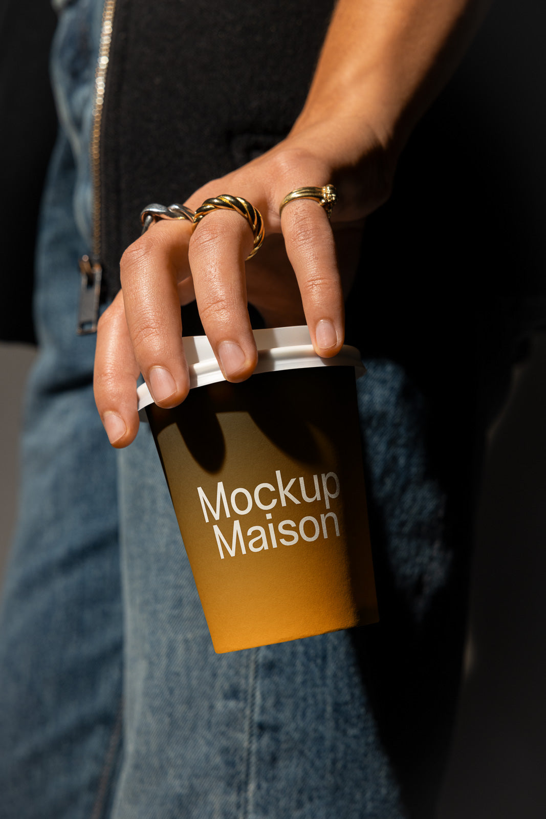 PSD mockup of a hand holding a brown paper coffee cup with the text "Mockup Maison," wearing gold and silver rings, with a denim and black jacket background.
