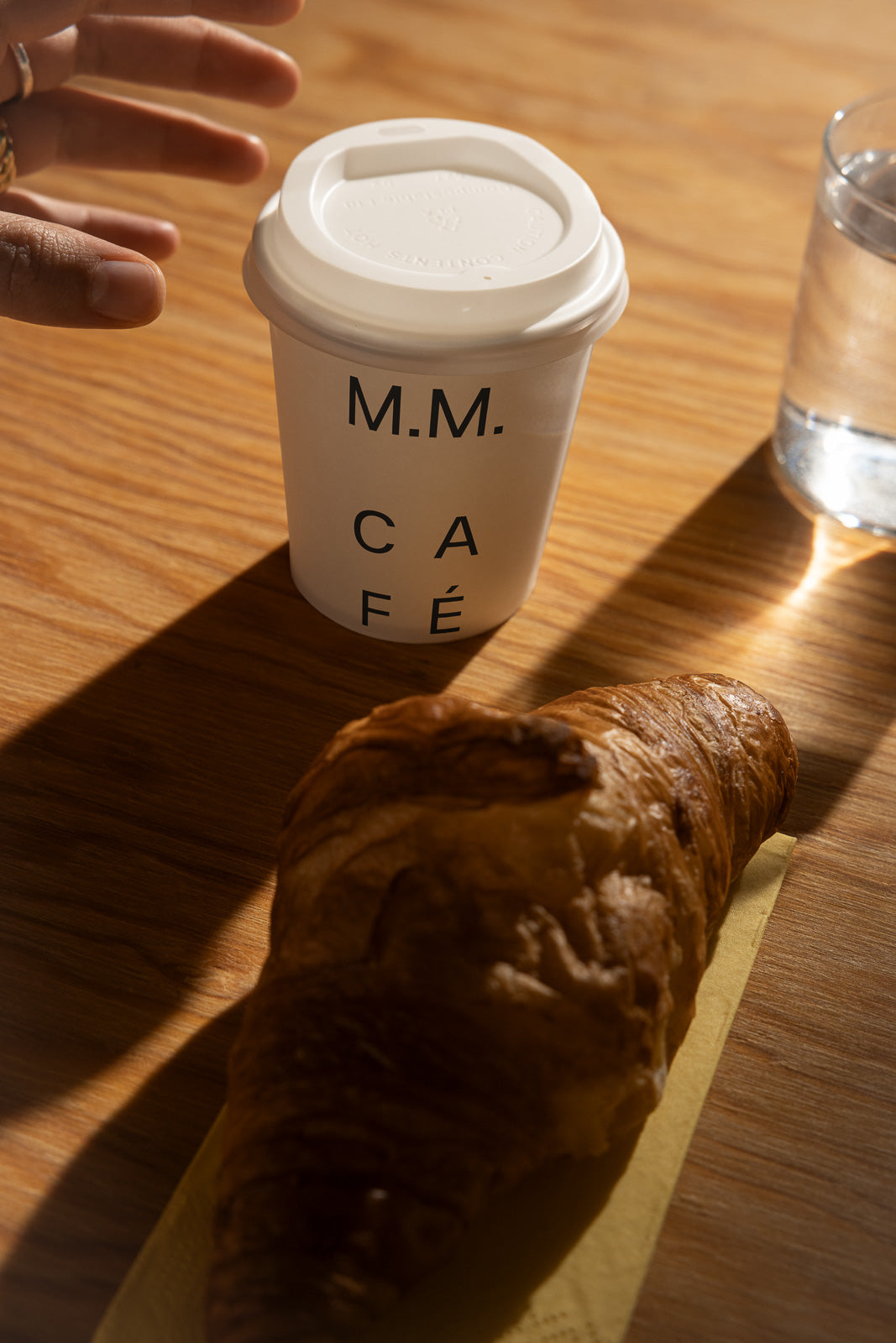 PSD mockup of a coffee cup labeled "M.M. Café" on a wooden table with a croissant and a glass of water nearby, sunlight casting soft shadows.