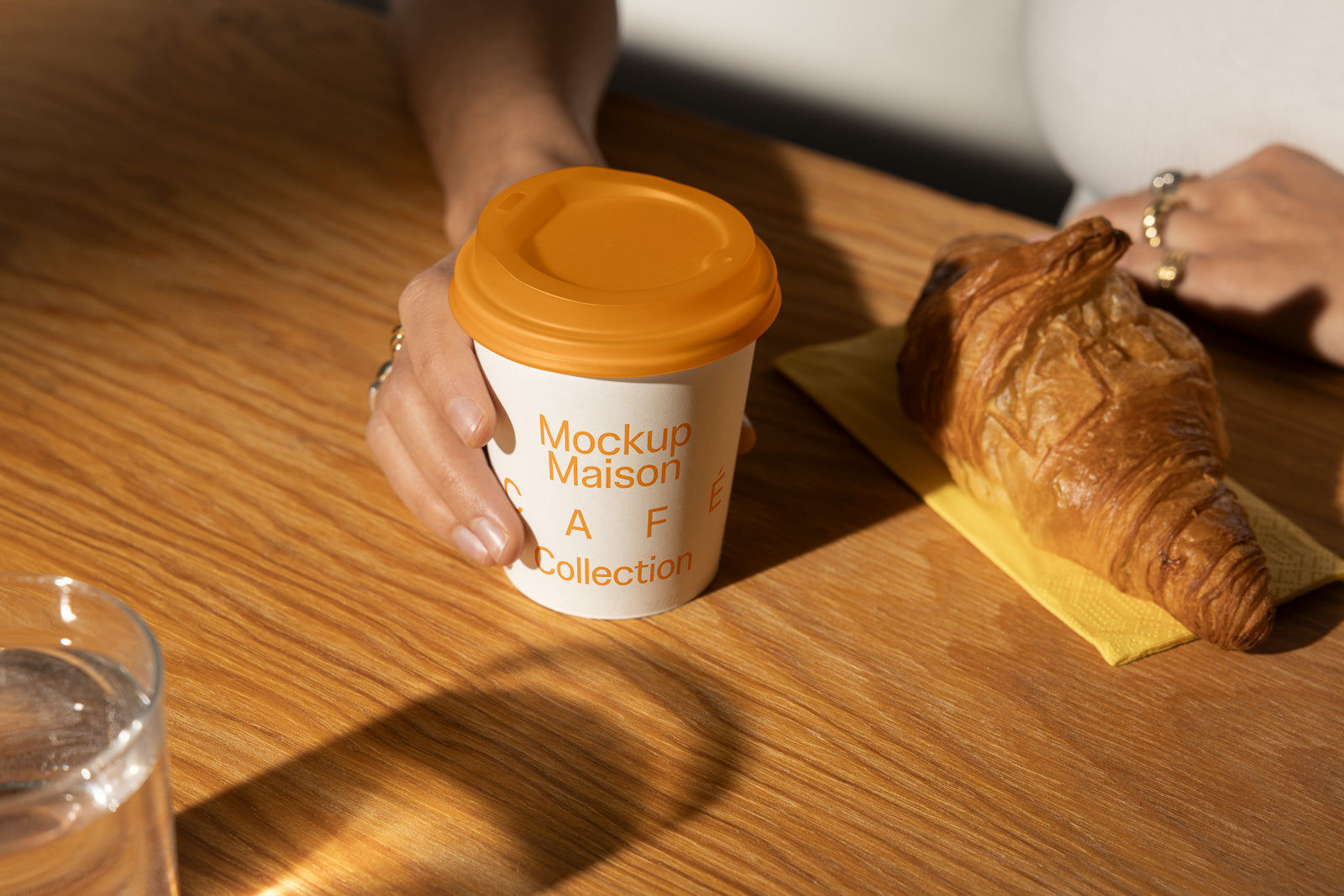 PSD mockup of a hand holding a paper coffee cup with orange lid on a wooden table beside a croissant and a glass of water.
