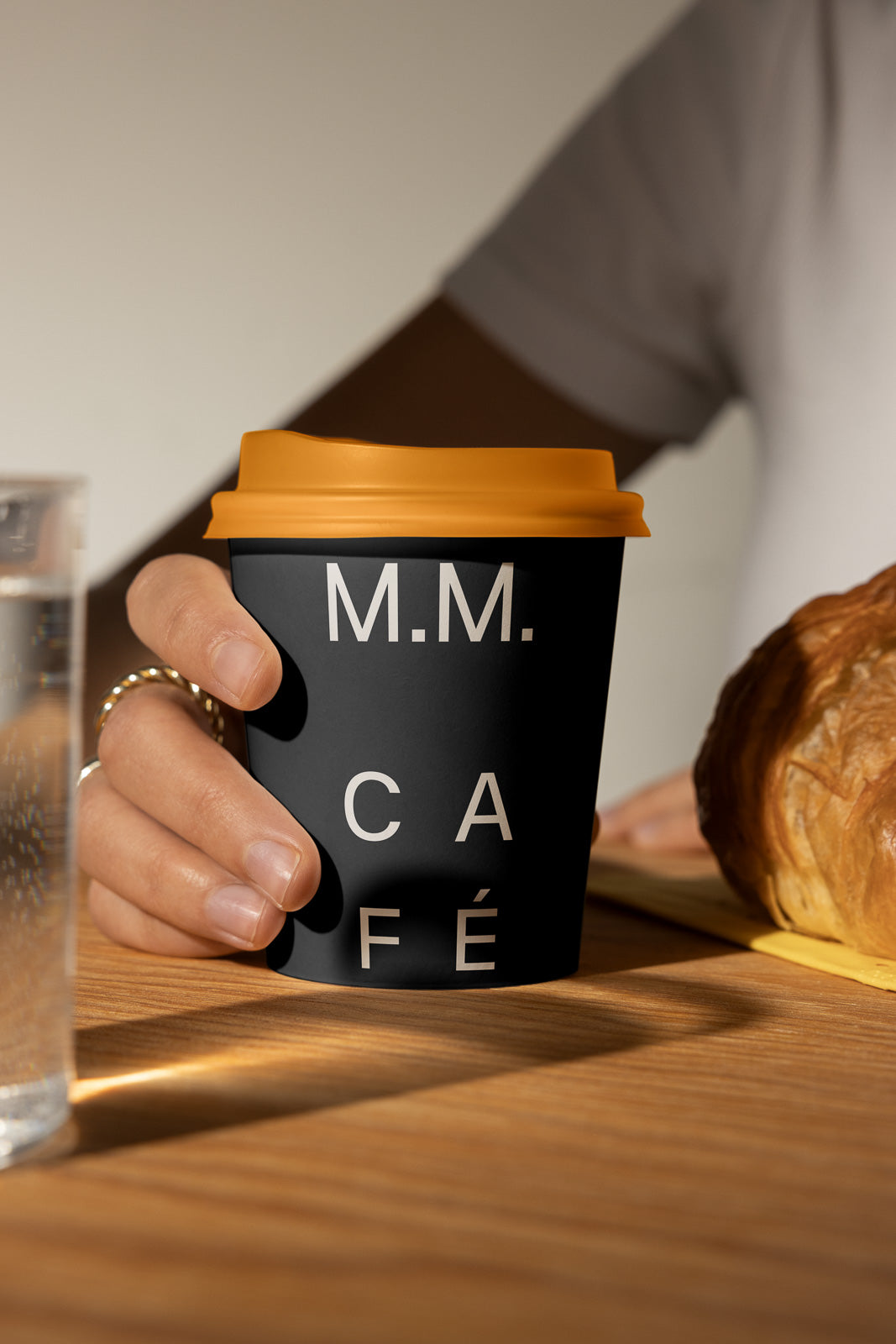 PSD mockup of a black coffee cup with "M.M. Café" in white letters and an orange lid, held by a hand on a wooden table near a croissant.