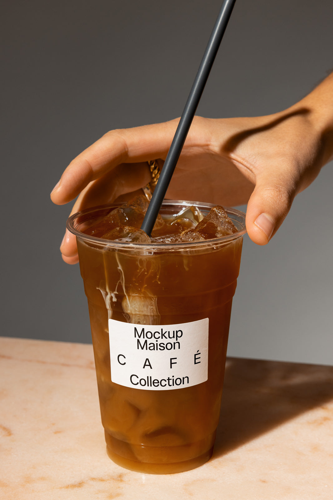 PSD mockup of a hand holding a plastic cup filled with iced coffee, labeled "Mockup Maison Café Collection," with a black straw inserted.