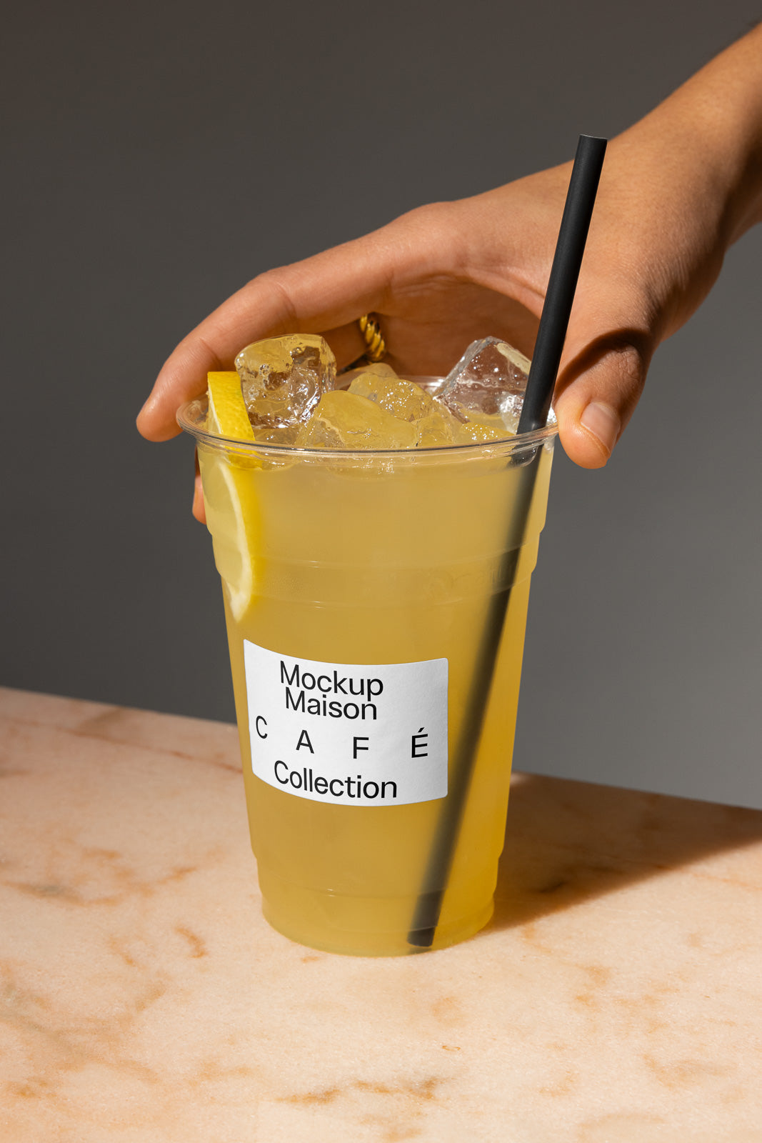 PSD mockup of a hand holding a plastic cup filled with iced lemonade, garnished with a lemon slice and a black straw. The label reads "Mockup Maison Café Collection."