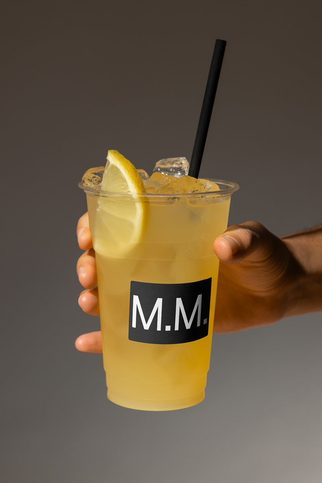 PSD mockup of a clear plastic cup filled with iced lemon drink, held by a hand. The cup features a black label with the letters "M.M." printed on it.