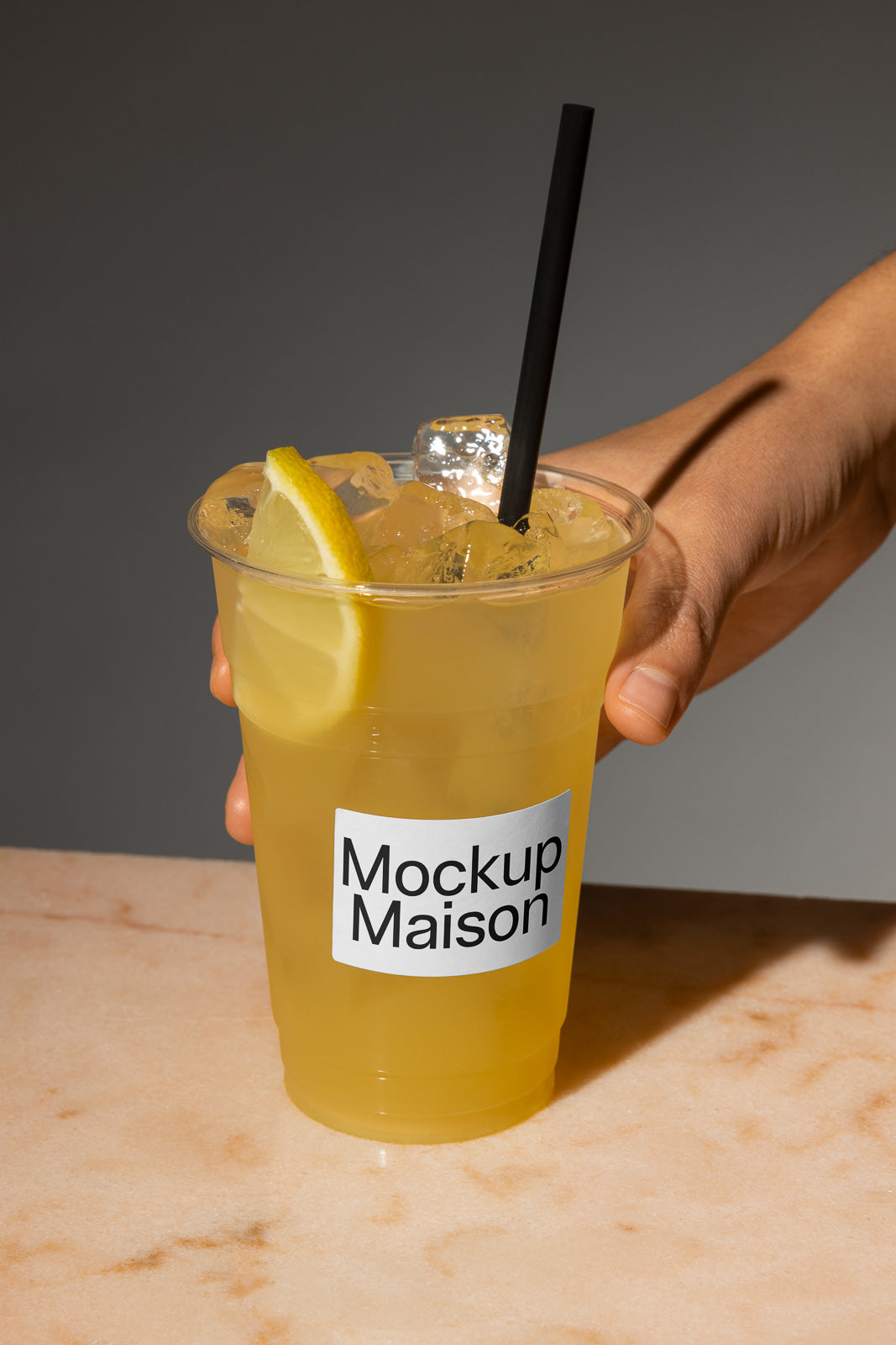 PSD mockup of a hand holding a clear cup filled with iced lemonade, labeled "Mockup Maison," on a marble surface, with a black straw and lemon slice.