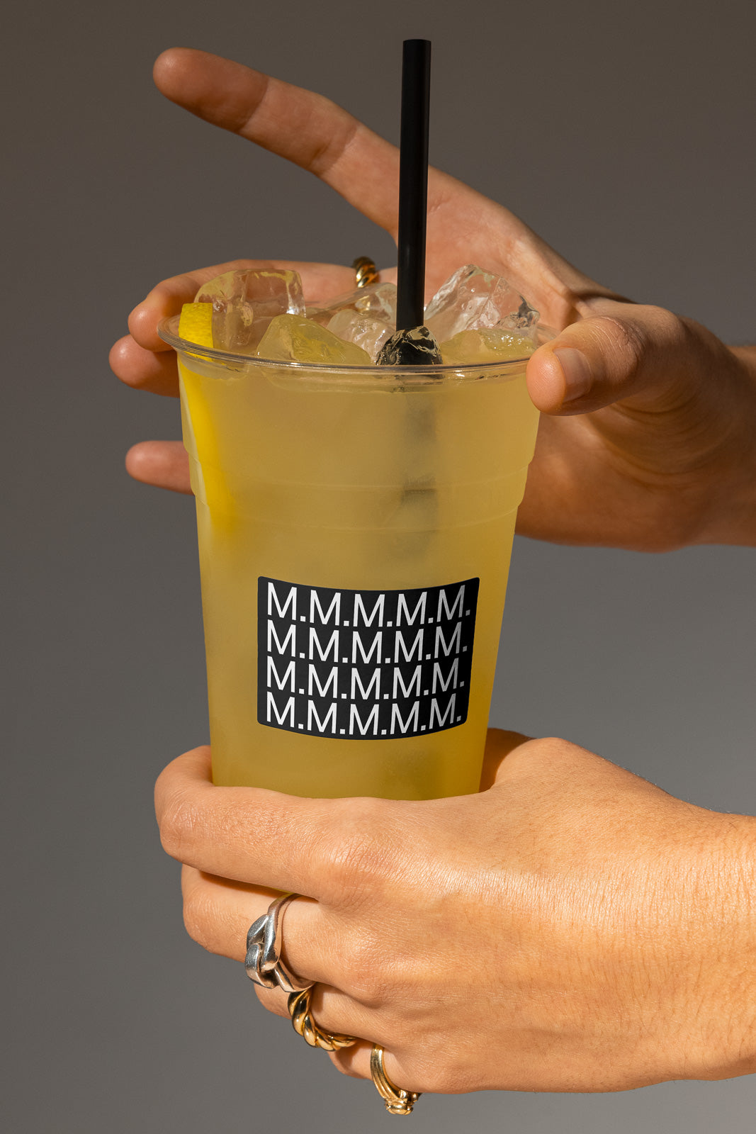 PSD mockup of a plastic cup with ice and lemon slices, held by a hand. The cup features a repeated "M.M.M.M." label and contains a black straw.